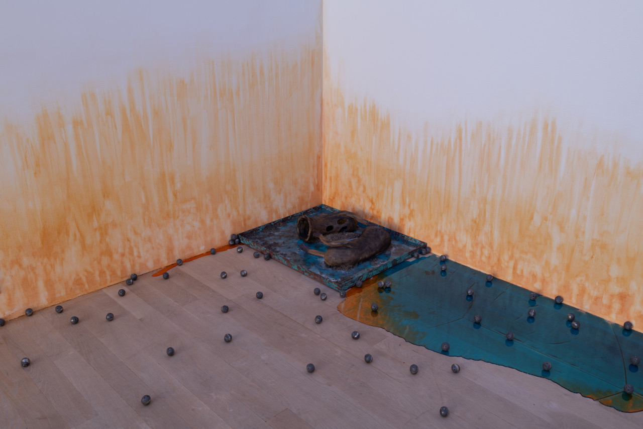 In a corner of the gallery, two oblong ceramic ghosts lay on their sides on top of a rusty blue square with small piles of charcoal. A shiny blue and orange pool of resin appears to leak out along the edges of the wall and numerous small metal balls are scattered around. On the wall, an orange stain appears to emerge from the floor.