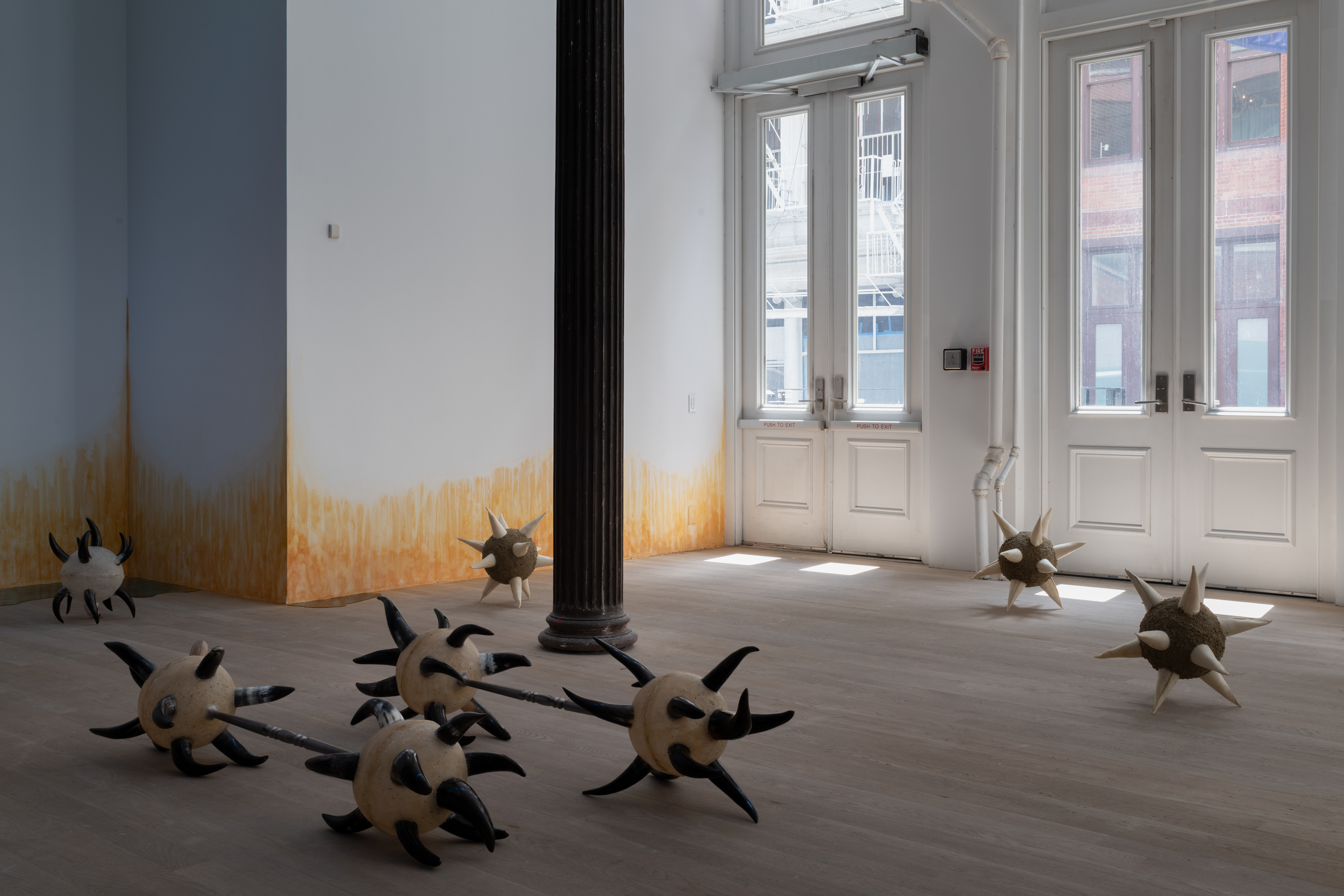 A sunlit gallery space is filled with four individual morning stars and two pairs connected by a chrome spindles, which are various shades of black, brown, and white. On the wall, an orange stain appears to emerge from the floor.