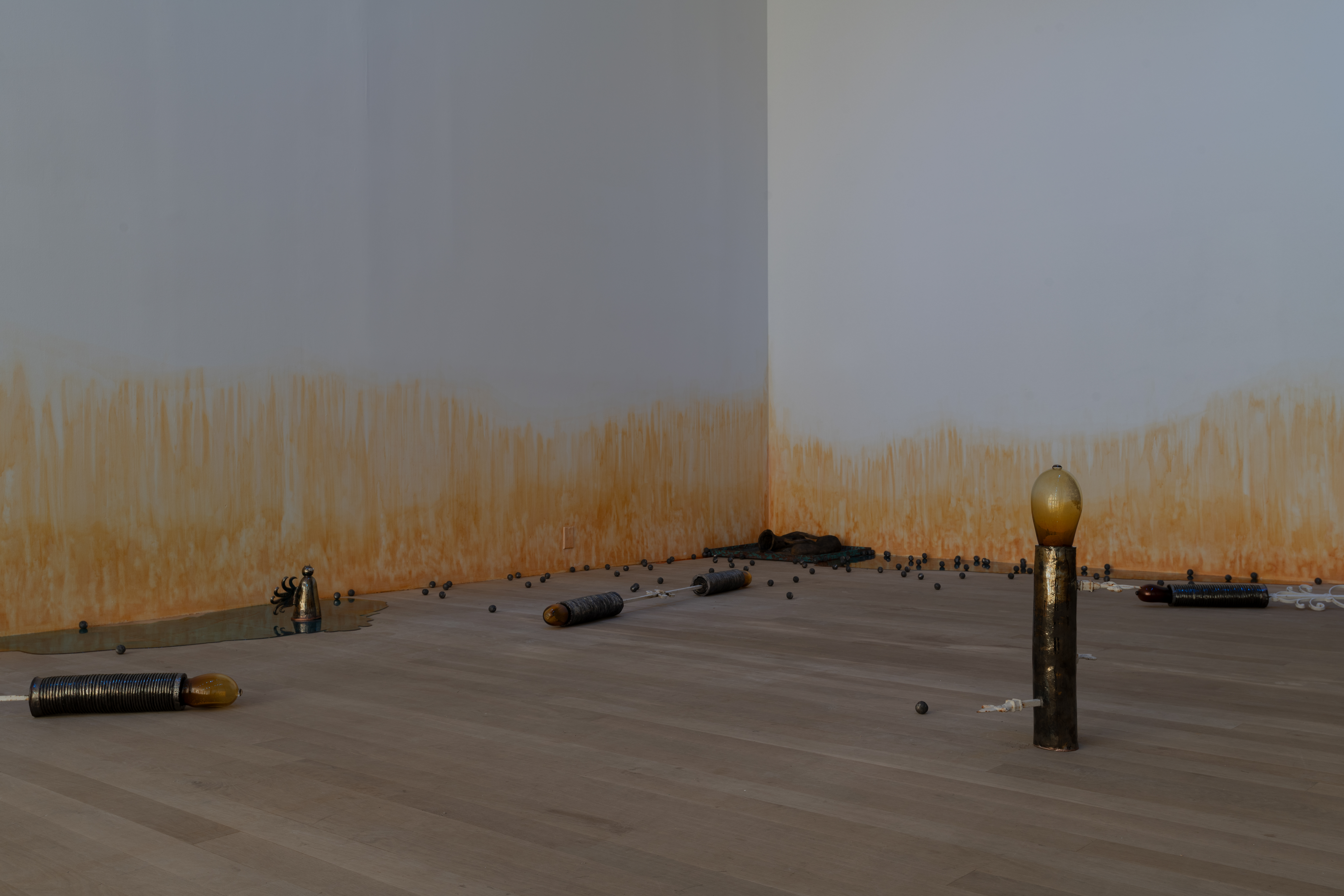 Four wrought iron sugar cane factory rollers, with amber-colored glass ovals at the ends, are arranged on their sides and upright on the floor. Behind, various other sculptures are seen, including metal balls, resin pools, oblong ceramic ghosts, and a small ceramic figure with horns and curly filamented wings. On the wall, an orange stain appears to emerge from the floor.