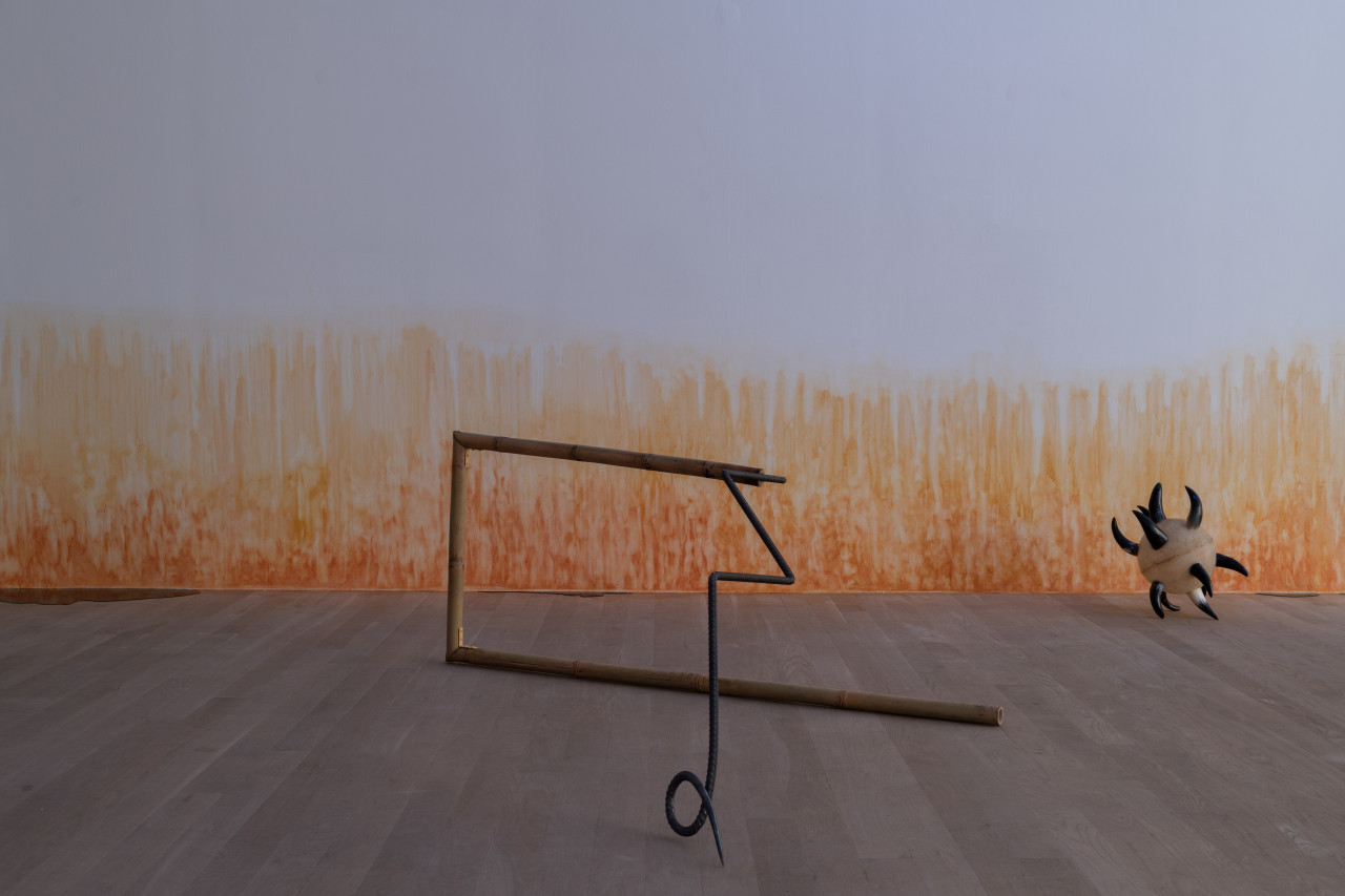 A bamboo and rebar spear that has been shaped into a rectangle is balanced on its side and curled tip. A single black and white morning star is arranged on the floor to the right. On the wall, an orange stain appears to emerge from the floor.
