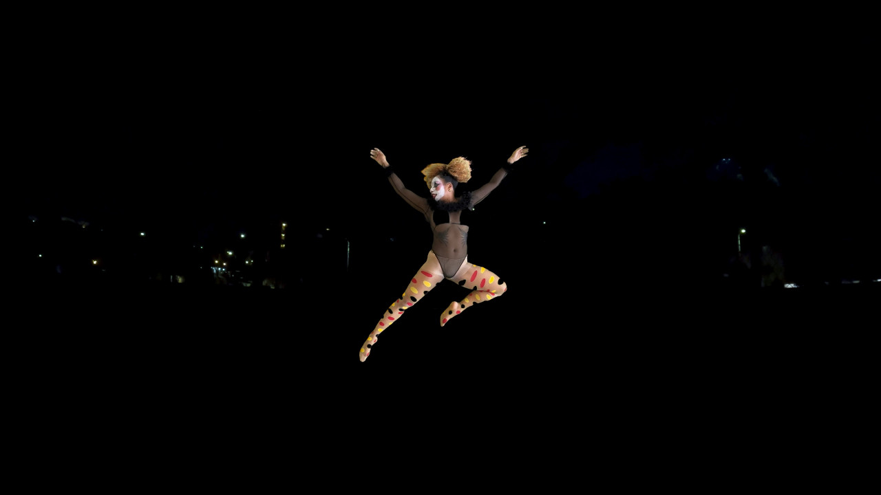 A figure in clown makeup, a black leotard, and polka dot tights jumps through the air against a dark background from which a few lights emerge.