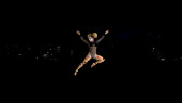 A figure in clown makeup, a black leotard, and polka dot tights jumps through the air against a dark background from which a few lights emerge.