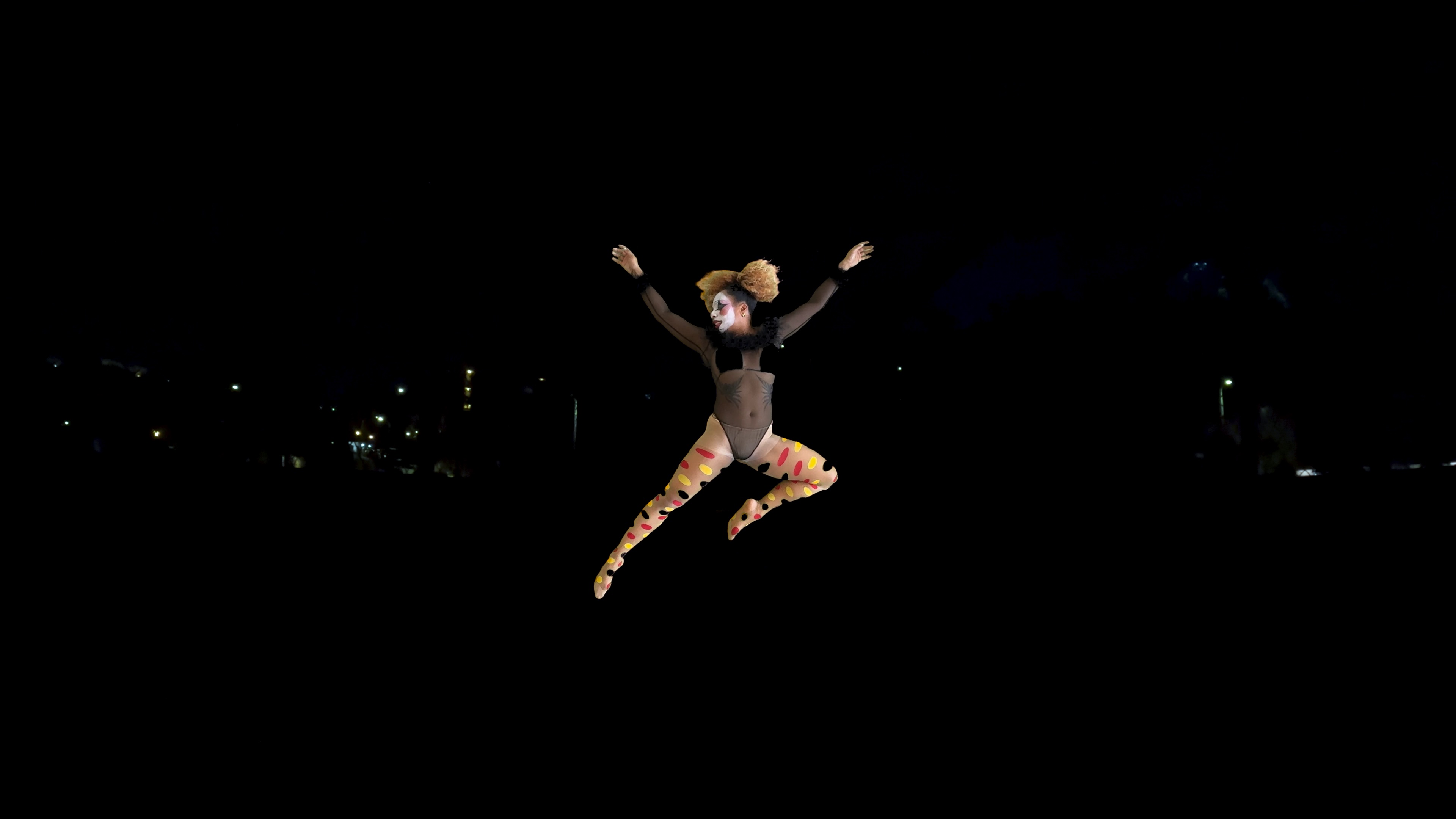 A figure in clown makeup, a black leotard, and polka dot tights jumps through the air against a dark background from which a few lights emerge.