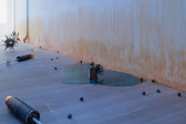 A small cylindrical ceramic figure, with curly filamented wings and a round head studded with horns and cowrie shell eyes, sits atop a reflective blue pool of resin with orange tints at the edges. Various other sculptures are are seen, including small metal balls, two morning stars, and two wrought iron sugar cane factory rollers with amber-colored glass ovals at the ends. On the wall, an orange stain appears to emerge from the floor.