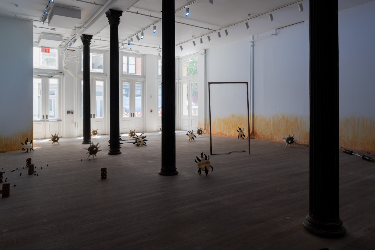 A sunlit gallery space is bisected by four columns through the middle. Various sculptures are arranged on the floor, including morning stars with cow horn spikes; a bamboo and rebar spear shaped into a rectangle; bricks wrapped in cowrie shells; resin pools; and ceramic balls. On the wall, an orange stain appears to emerge from the floor.