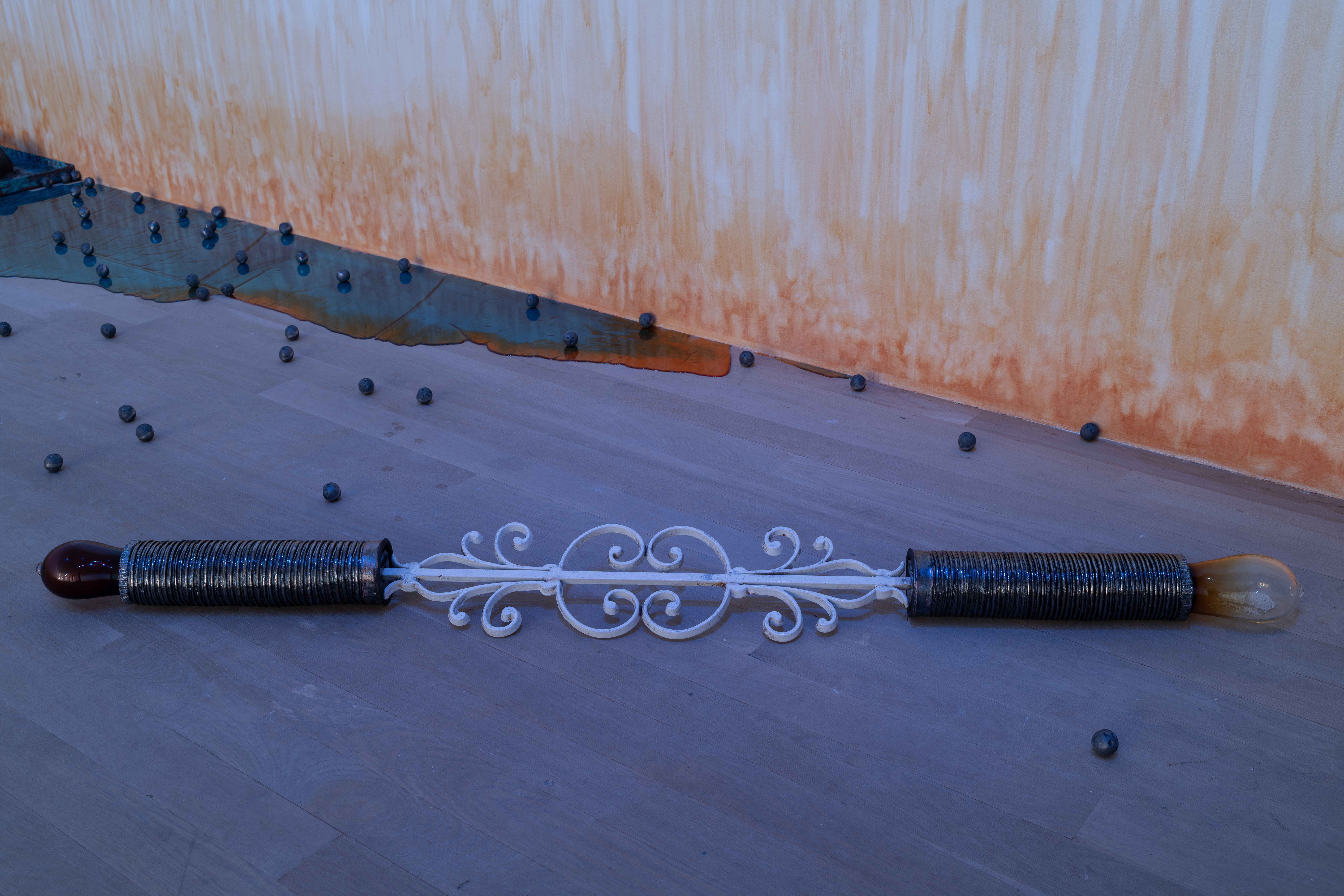 Two wrought iron sugar cane factory rollers with grooves and amber-colored glass ovals at the ends rests on the ground. They are connected by a white wrought iron rod in a spiral design. Behind, a shiny blue and orange resin pool along metal balls are arranged on the floor. On the wall, an orange stain appears to emerge from the floor.