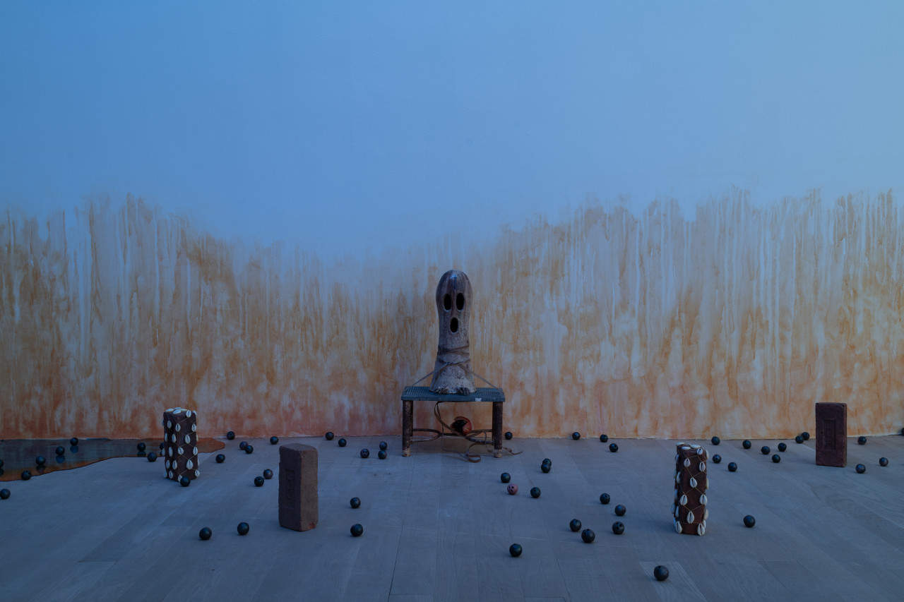 Along the orange-stained gallery wall, an oblong ceramic sculpture of a ghost with its mouth open is tied down to a metal stool with an ackee underneath. Four bricks are arranged around and two are wrapped in nets dotted with cowrie shells. Small ceramic balls are scattered on the floor, and a shiny blue and orange resin pool appears to leak from the left side of the wall.