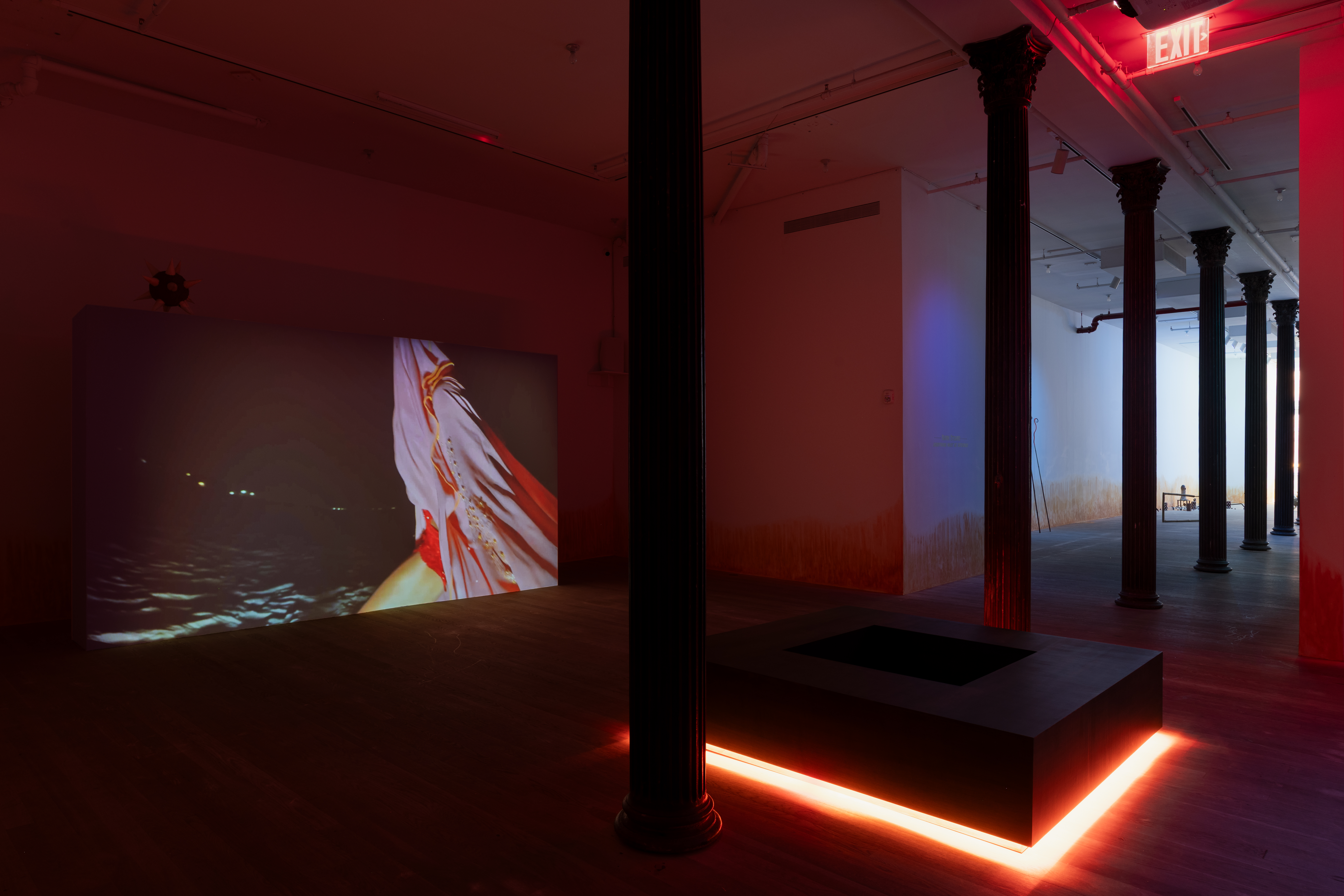 On the right between two columns in a darkened room, a square-shaped bench radiates bright orange light from below. On the left, a large projection on the wall shows a figure, whose head and body is draped in lilac and red fabrics, in motion on the beach at night.