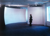 A color installation photograph of a figure in front of a curved white wall in a gallery space.