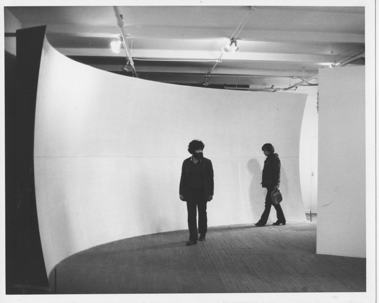 A black and white installation photograph of two figures in front of a curved white wall in a gallery space.