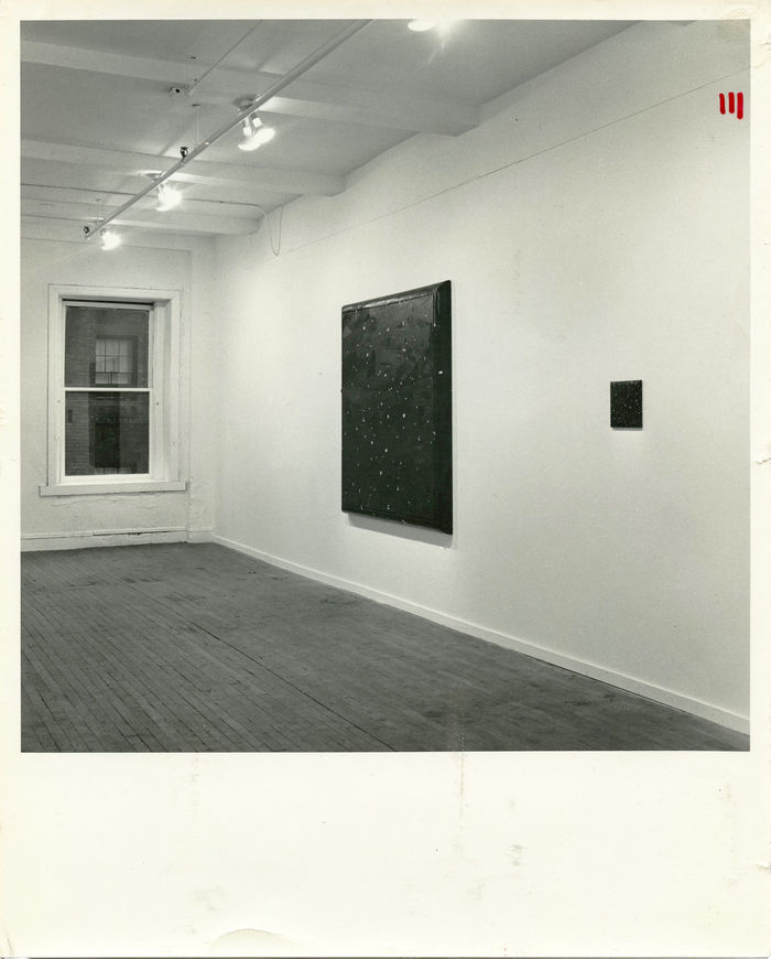 A black and white installation photograph of a gallery space. Two dark square canvasses hang on the wall, one is a large and the other small.