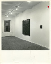 A black and white installation photograph of a gallery space. Two dark square canvasses hang on the wall, one is a large and the other small.