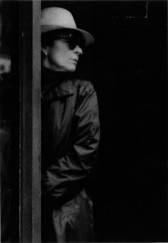 A person in a long coat, hat, and sunglasses leans against a doorframe.