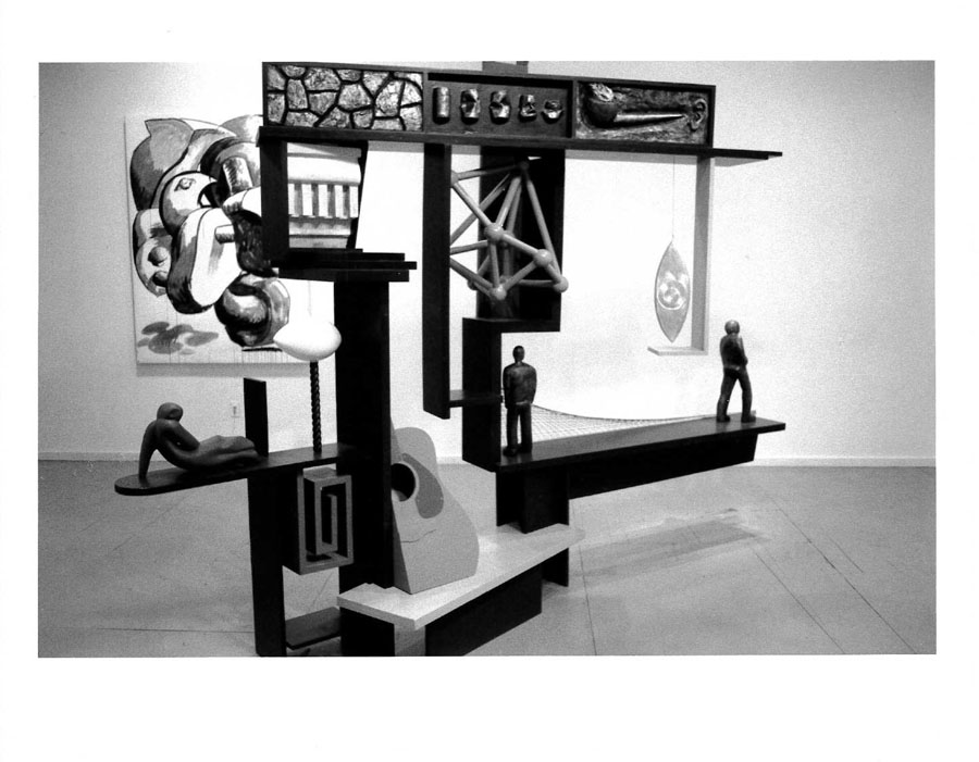 A number of small figurines appear on a sculpted object with various other items including a half of a guitar and a picture. All appearing in a white-walled gallery space.