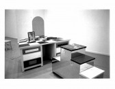 In the foreground lies a silver sculpture with varying levels and in the background is a table with a checkered design on top. An image stands on that table and a stool can be seen behind it.