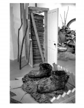 Sculpted boots appear wih leaves on a leaf-covered section of the floor surrounded by a picket fence. In the background a white door opens up with a stack behind it. Nearby tree branches and other rustic constructions populate the gallery space.