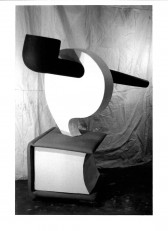 A black and white photograph of a black pipe sticking through a large white shape coming from a large book. A wrinkled curtain is visible behind the construction.