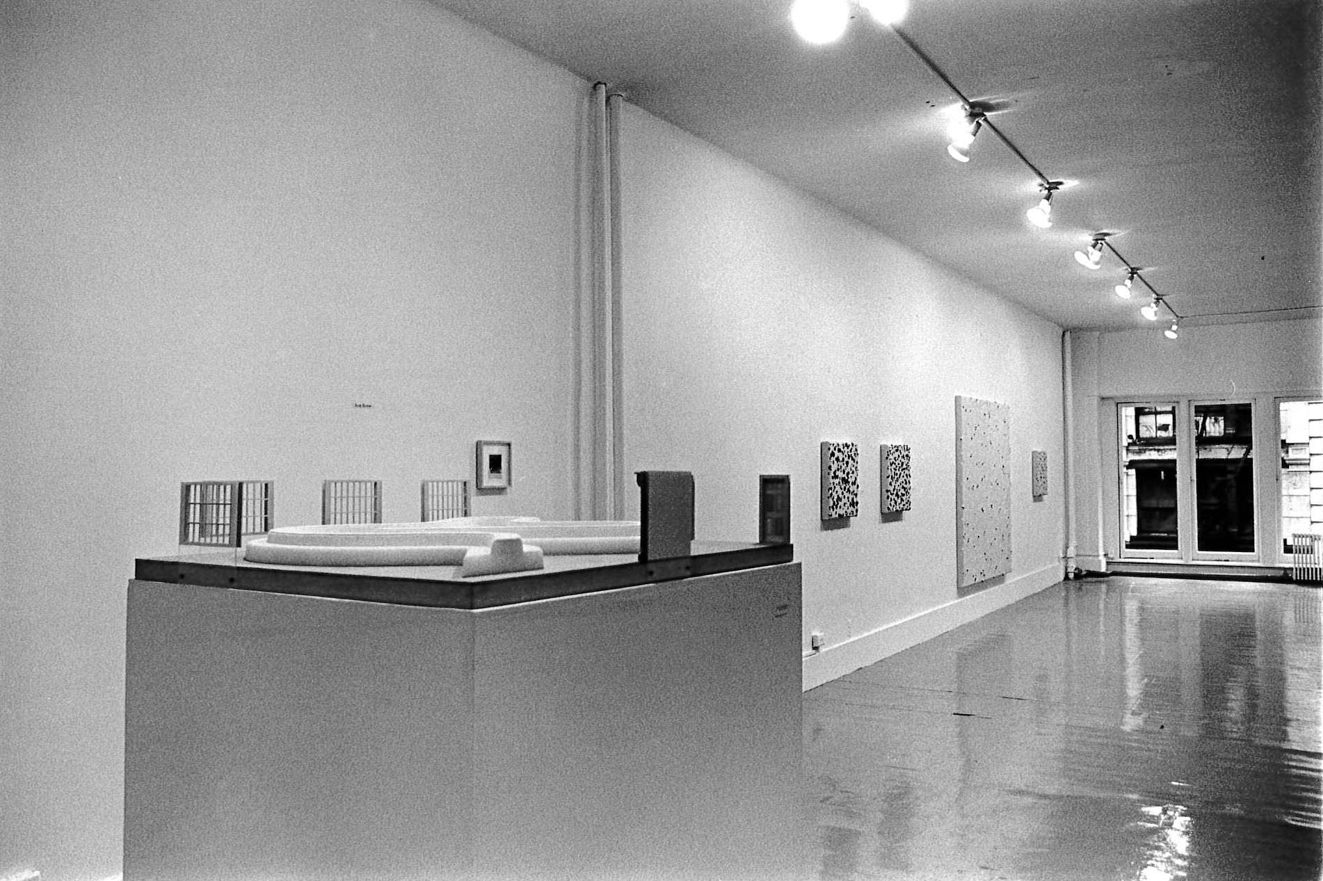In the foreground, a podium displays a curvy white object. Miniature doors and windows are attached to the top edges of the podium facing inward towards the tube-like white object.