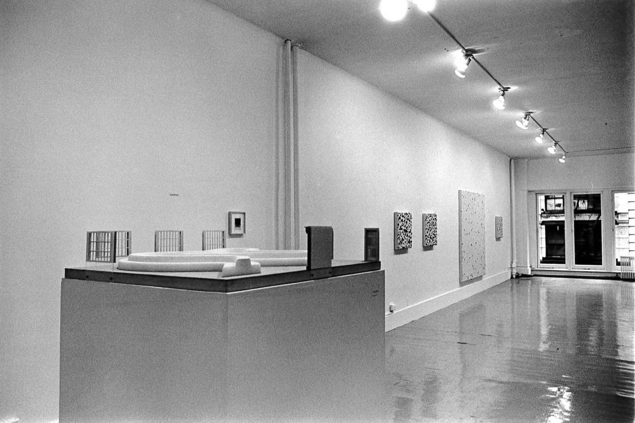 In the foreground, a podium displays a curvy white object. Miniature doors and windows are attached to the top edges of the podium facing inward towards the tube-like white object.