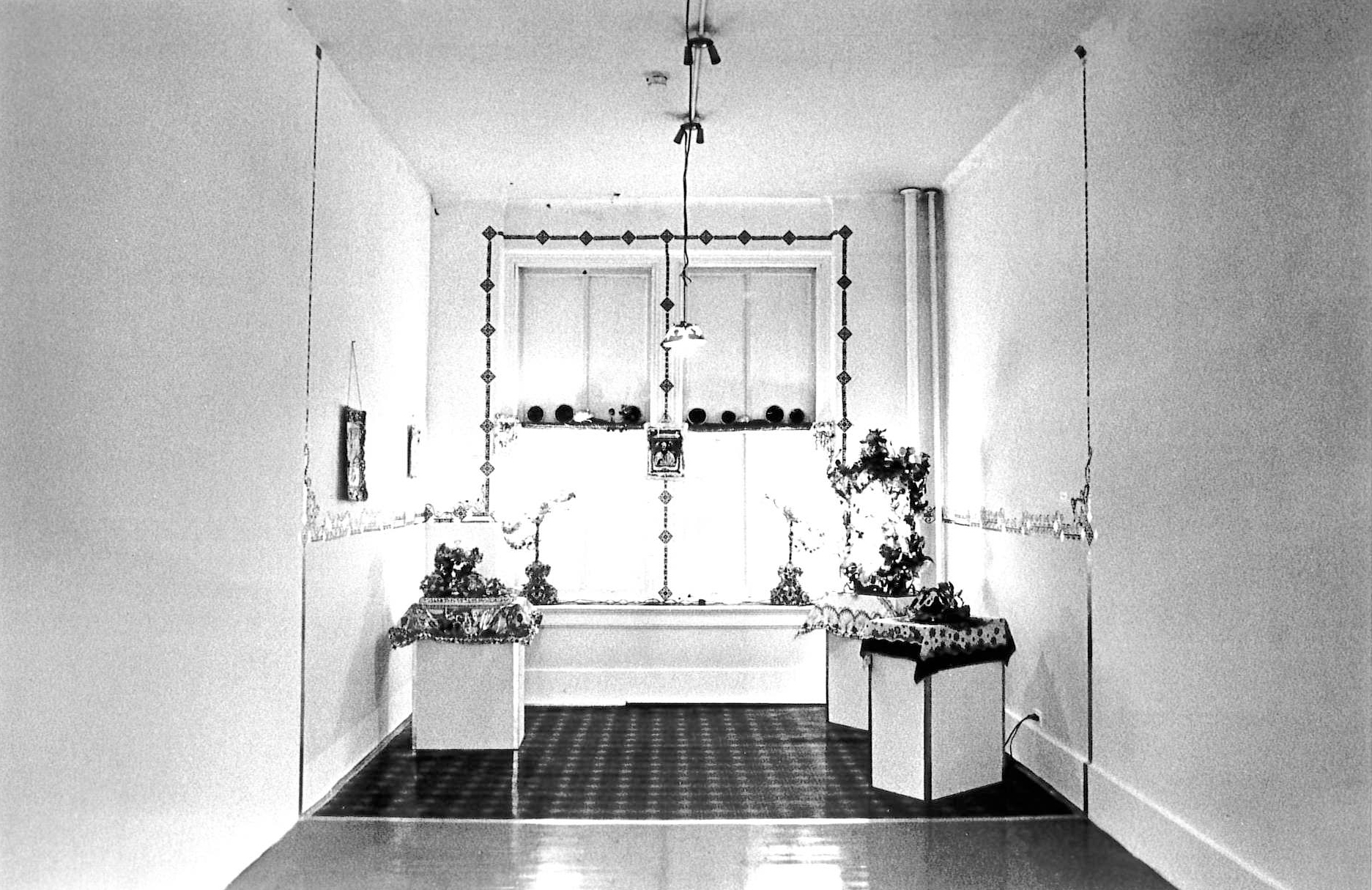 Three pedestals, covered with fabric scarves and various objects, sit atop a dark floor covering. Framed images hang on the walls. Along the back wall, large windows are adorned with candlesticks.