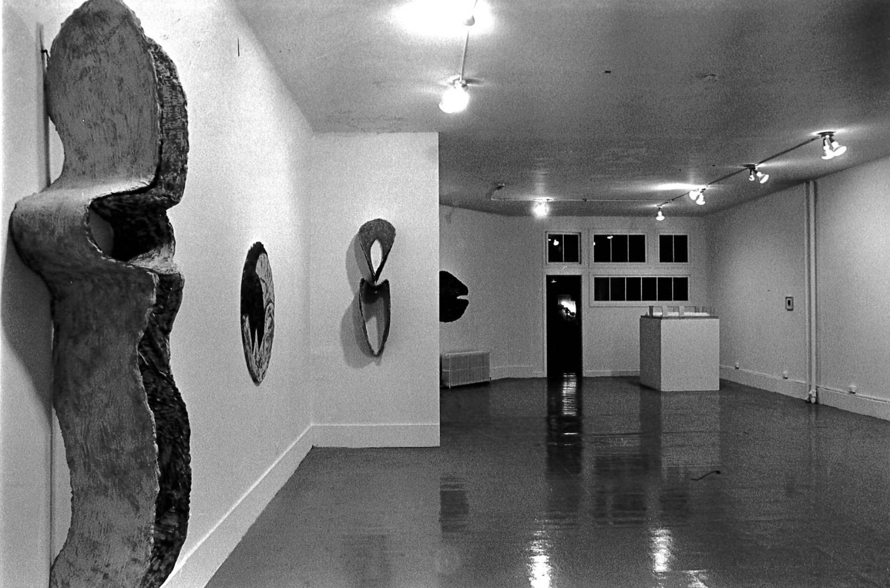 Sculptural pieces mounted to the left wall. A white display pedestal sits in the background on the right, next to an open door.
