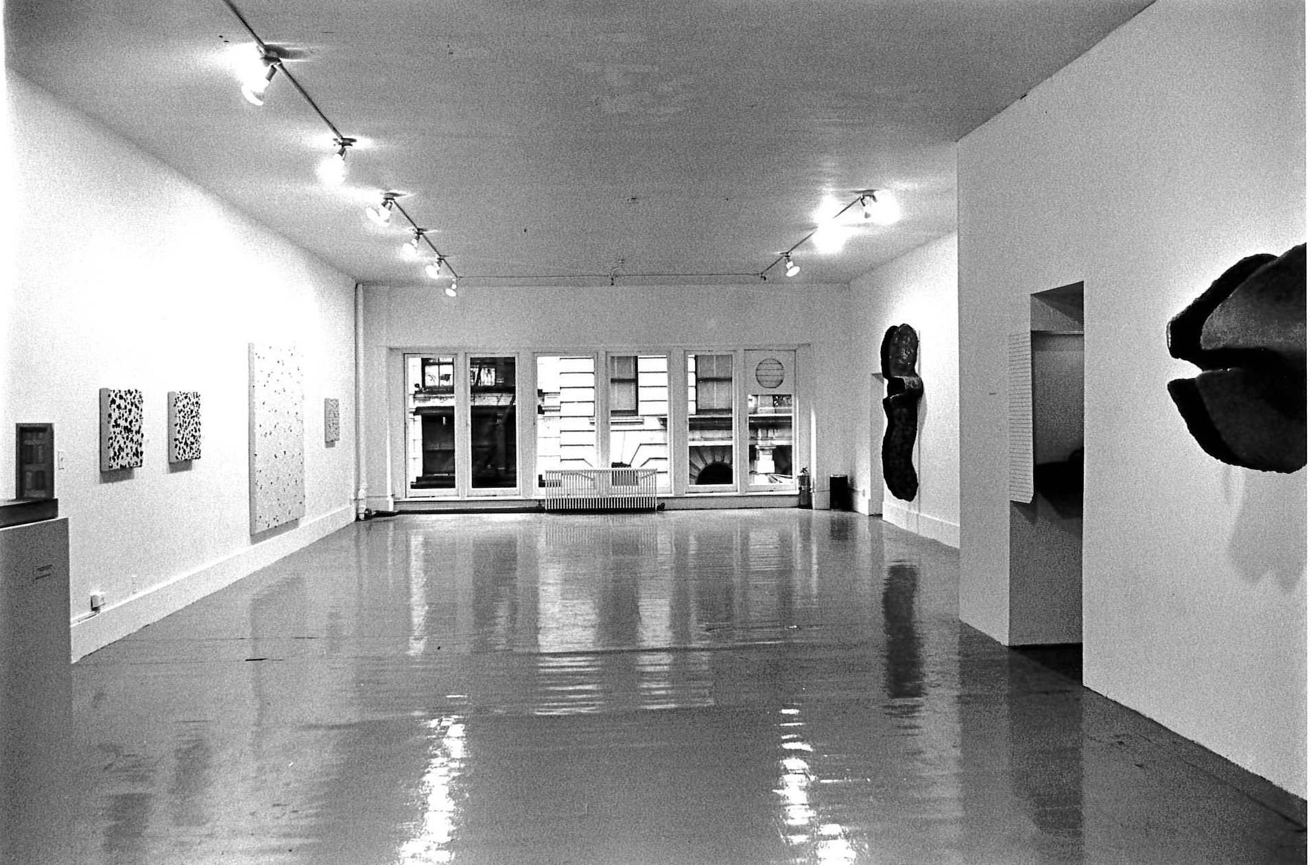 Sculptural pieces are mounted on the right wall. Three square canvases and one larger rectangular canvas dotted with a dark material hang on the left wall. Large windows line the back wall.