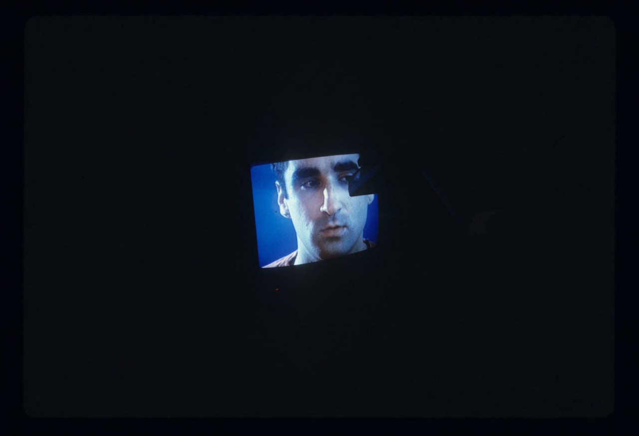 A color television CRT monitor is visible with a man's face on it. This monitor is shrouded in darkness and some indistinguishable object appears to be touching the screen.