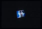 A color television CRT monitor is visible with a man's face on it. This monitor is shrouded in darkness and some indistinguishable object appears to be touching the screen.