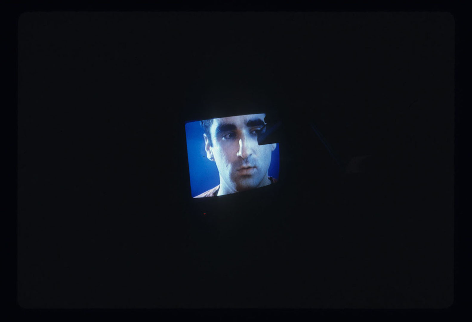 A color television CRT monitor is visible with a man's face on it. This monitor is shrouded in darkness and some indistinguishable object appears to be touching the screen.