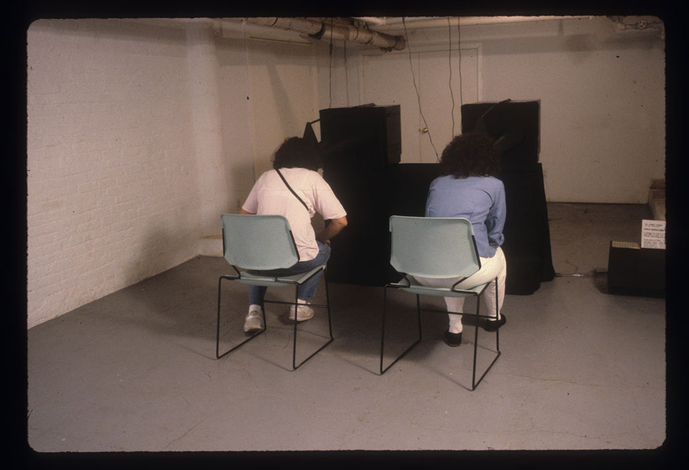 Two people sit on chairs in front of television monitors that are showing a black image. The television monitors are on pedestals in the gallery space.