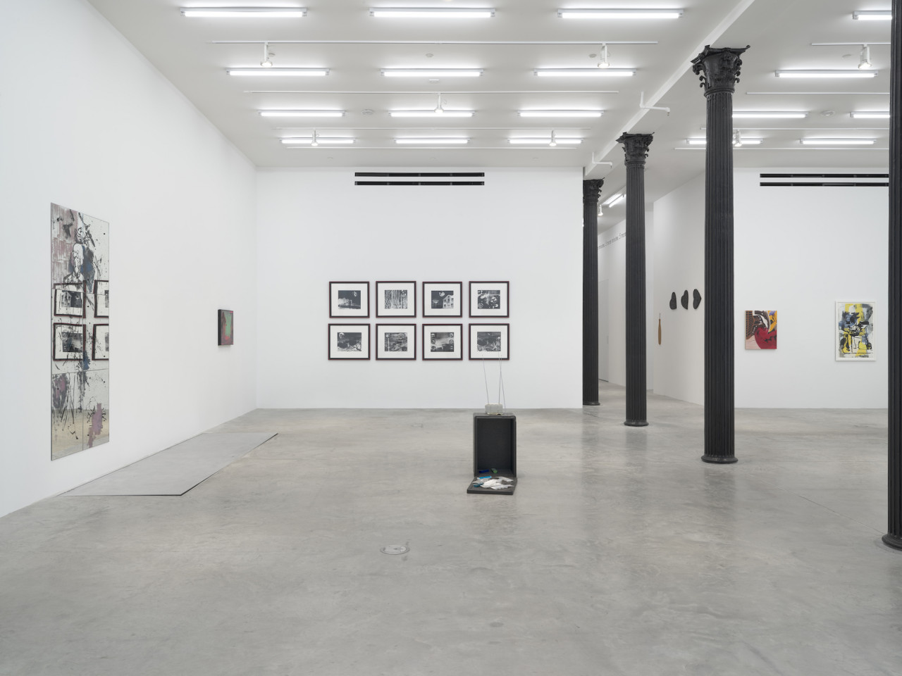 A view of paintings and photographs across three walls in the space. Black columns divide the room; to the left, a sculpture is displayed on the ground.