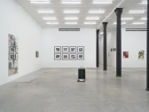 A view of paintings and photographs across three walls in the space. Black columns divide the room; to the left, a sculpture is displayed on the ground.