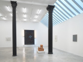 A large painting illuminated by lights hangs on a wall behind two black columns. A small sculpture sits on the ground in front of it. Smaller artworks are visible on the connecting walls.