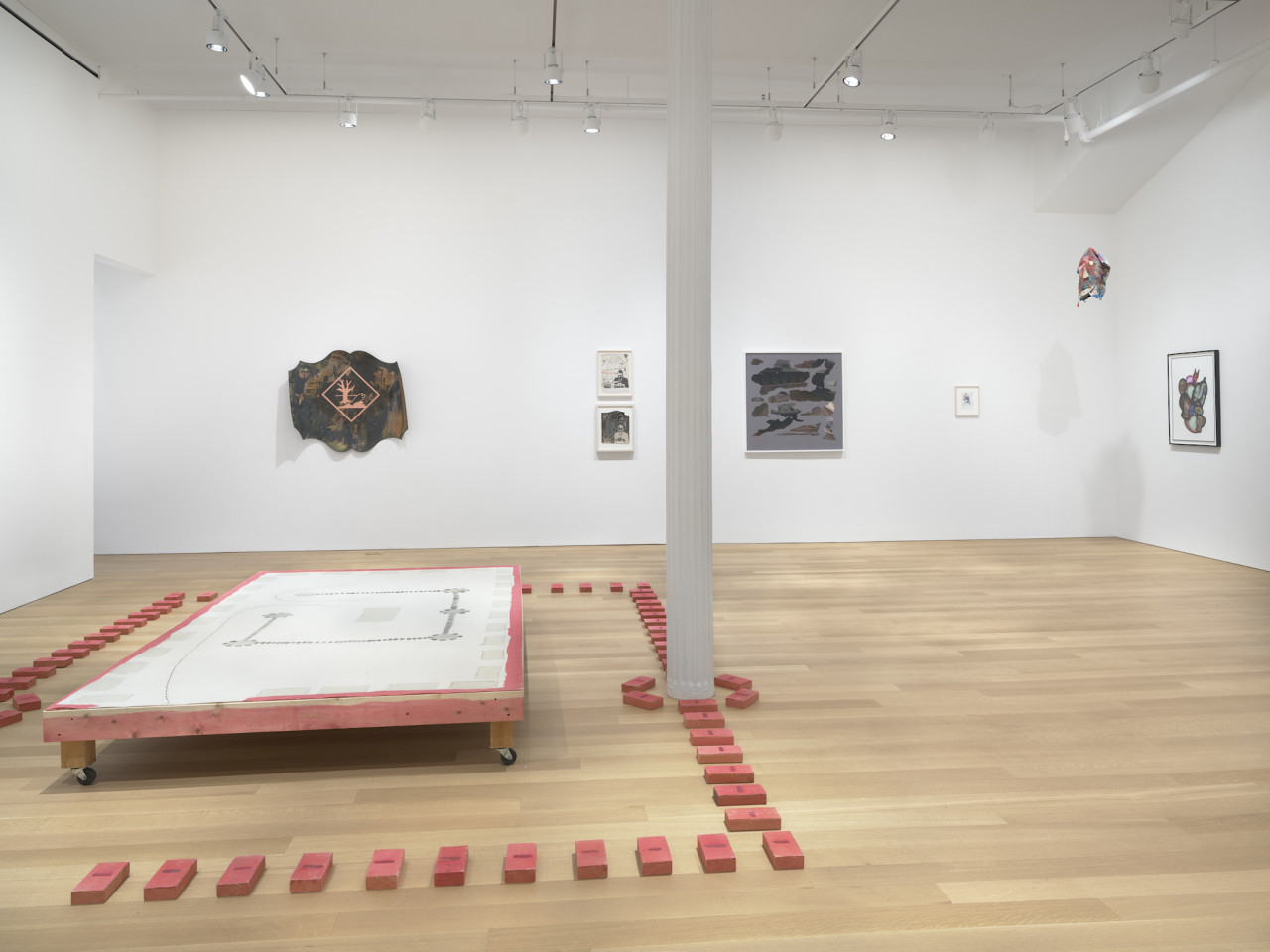 A wooden platform on wheels surrounded by a border of small poppy red brick-like sculptures, that continue to surround the base of a white pole. Sculptures and paintings are visible on the back wall behind the wooden platform.