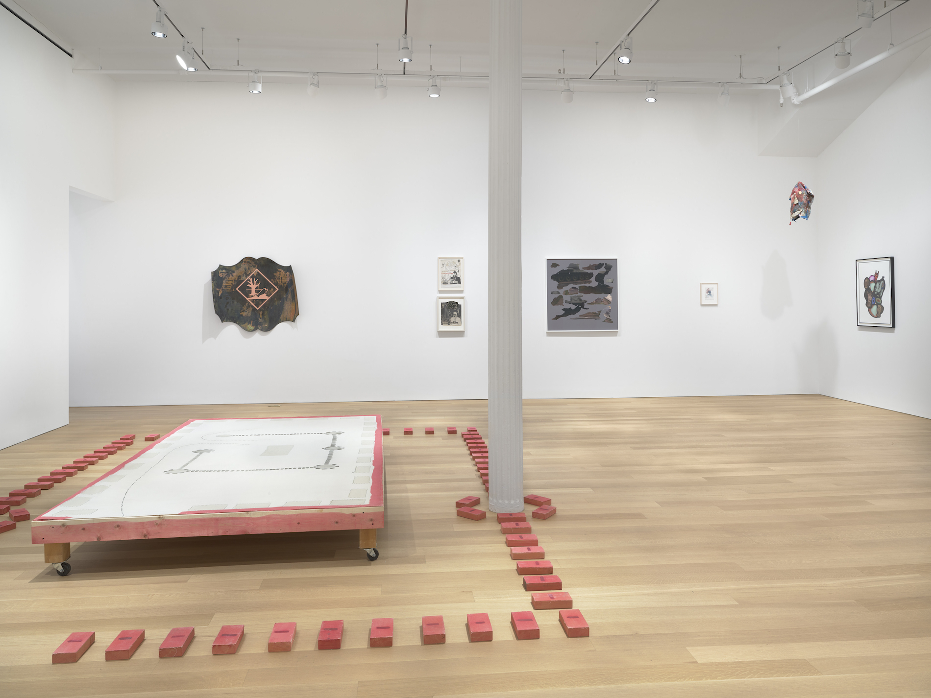 A wooden platform on wheels surrounded by a border of small poppy red brick-like sculptures, that continue to surround the base of a white pole. Sculptures and paintings are visible on the back wall behind the wooden platform.