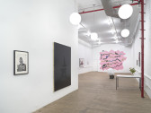 An angled view of paintings and photographs against multiple walls. In the background, a pink wall installation is mounted.