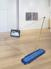 A wide blue mop with a yellow handle is tethered to a white pillar by a black cord, and is stretched with the handle angled forward. Resting against the pillar is a tv, angled and facing slightly leftwards, displaying a sculpture in a parking lot.