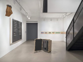 An opened case, with camera equipment resting inside gray foam, is displayed in the center of a room. A large black and white painting with text is hung on a wall underneath a hanging sculpture. In the background, individual papers of beer logos are displayed behind glass, stretching across two connecting walls.