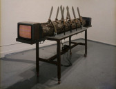 A large log spans the length of a table, held down with straps and with eight machetes affixed to the table alongside it.  On either end of the log are two televisions, and the one facing the camera displays static.