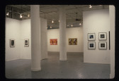 Framed square and rectangualar photographs are visible in a white-walled gallery space with white columns centered in the image.