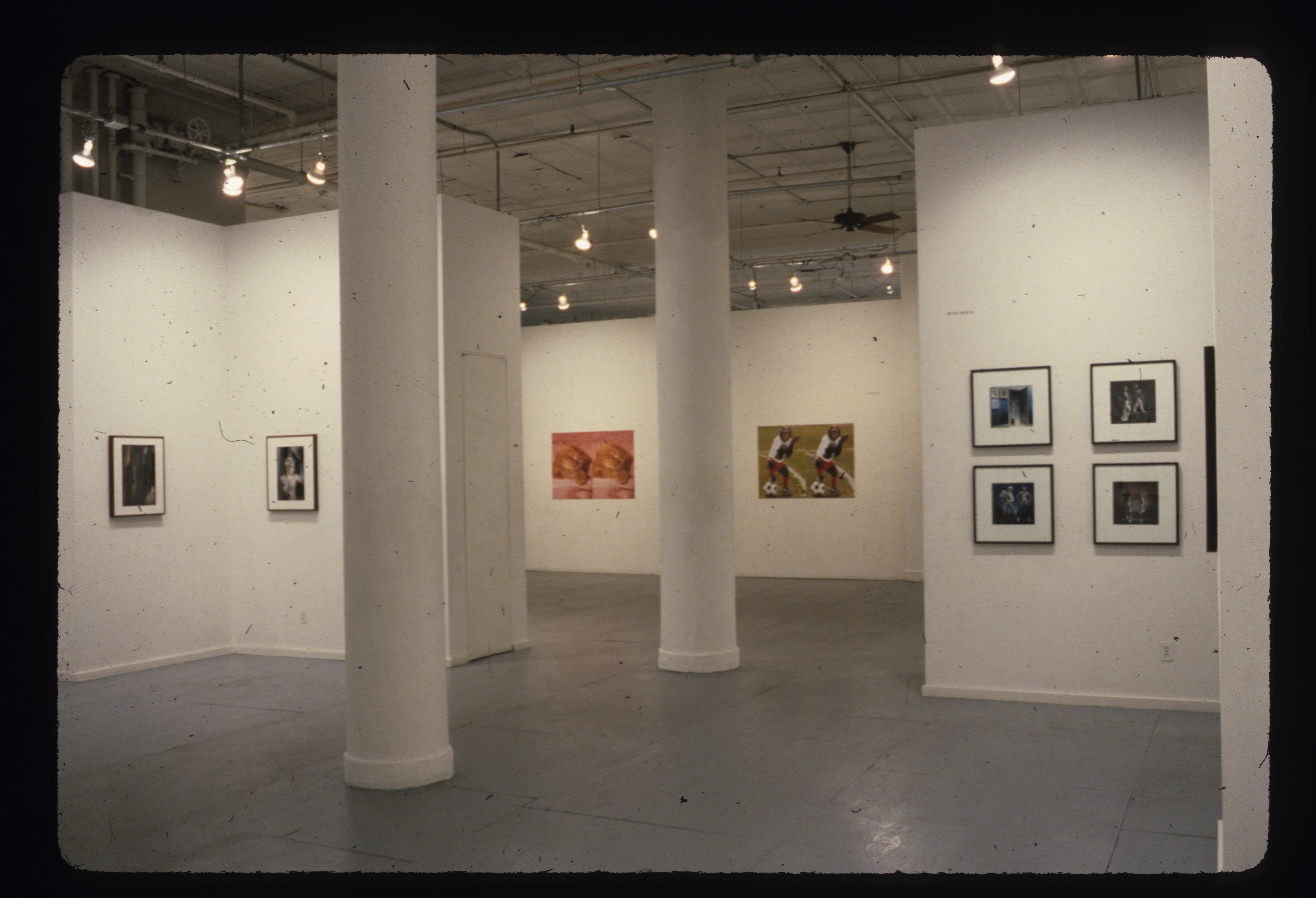 Framed square and rectangualar photographs are visible in a white-walled gallery space with white columns centered in the image.