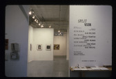 Papers lies on a table in front of exhibition wall text, baskets with paper are hanging on an adjacent wall. Framed paintings can be seen in the white-walled gallery space in the background.