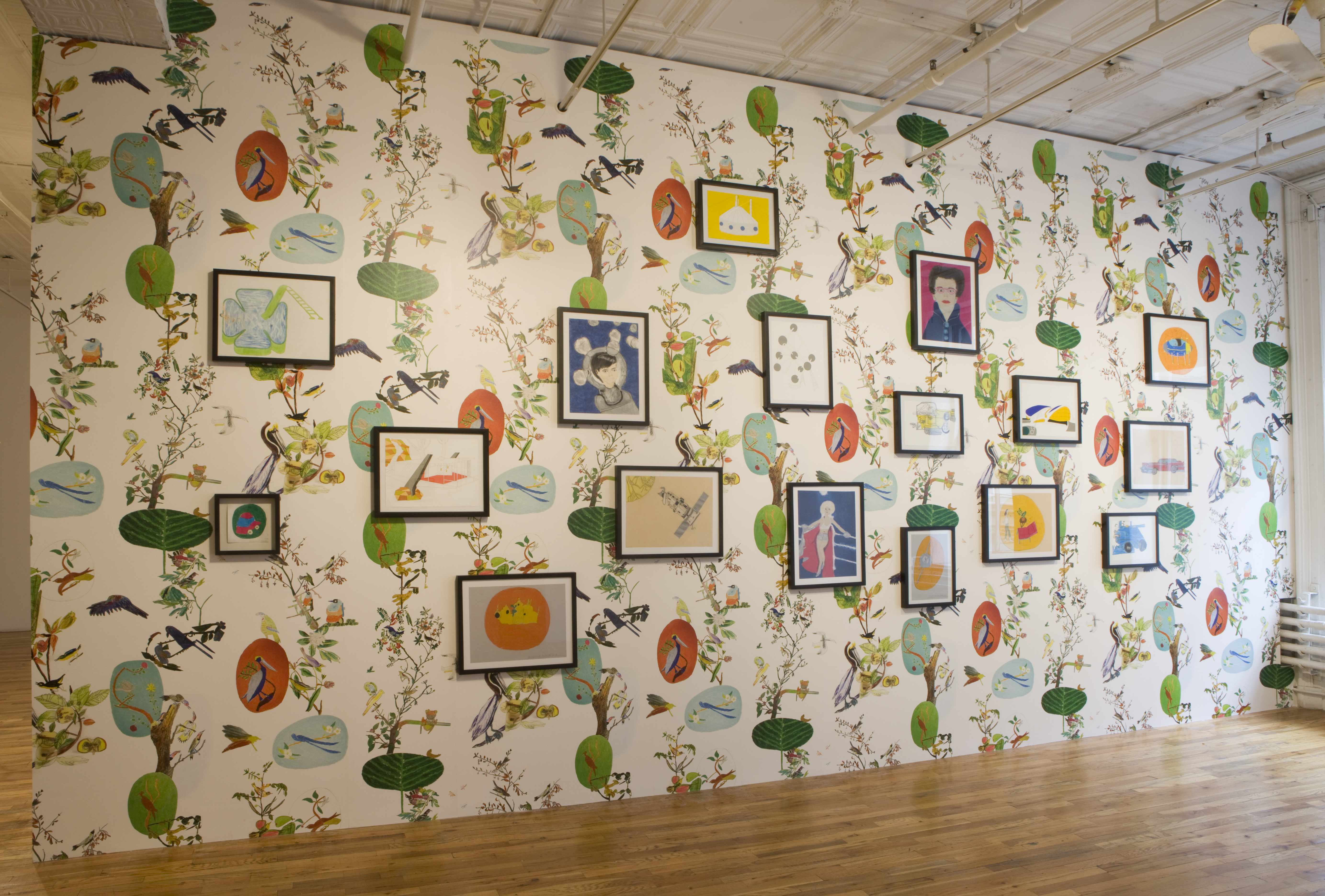 A large gallery wall covered with wallpaper printed with tropical birds in colorful trees on a white background. There are assorted black-framed, colorful images hung on the wall, positioned in both portrait and landscape orientations.