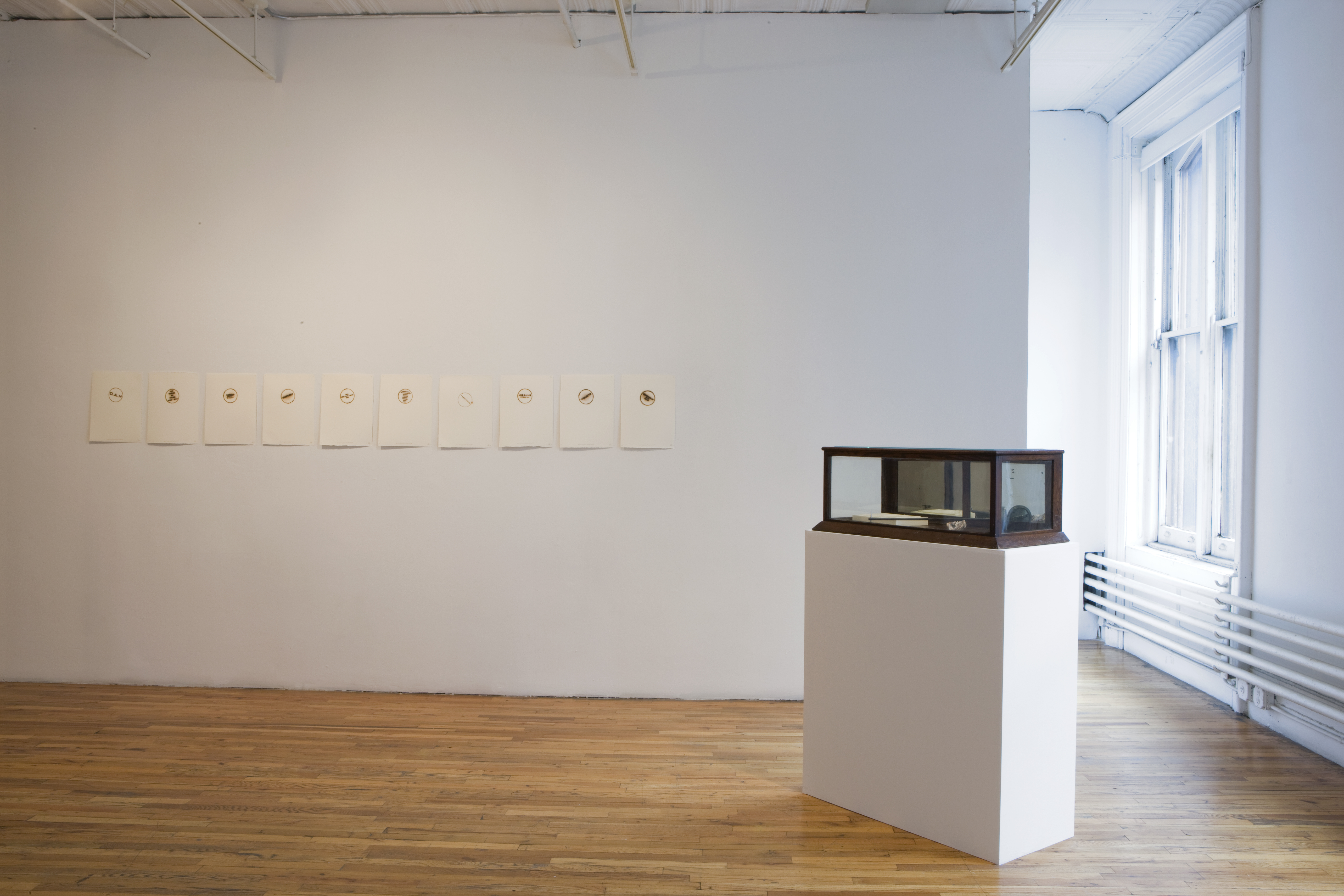 Ten white pieces of paper hang on a wall, each with a slightly varying circular brand. To the right of the wall is a white plinth that supports a wooden and glass case that houses several objects.