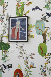 A painting of a woman with blonde hair and a cape hangs on a wallpapered wall. The wallpaper is white and decorated with colorful images of tropical birds in trees.