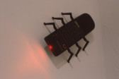 A black remote control with a red light pointing out of its head. Six insect-like legs are attached to the body of the remote, three on each side, and it is poised on the wall as if crawling.