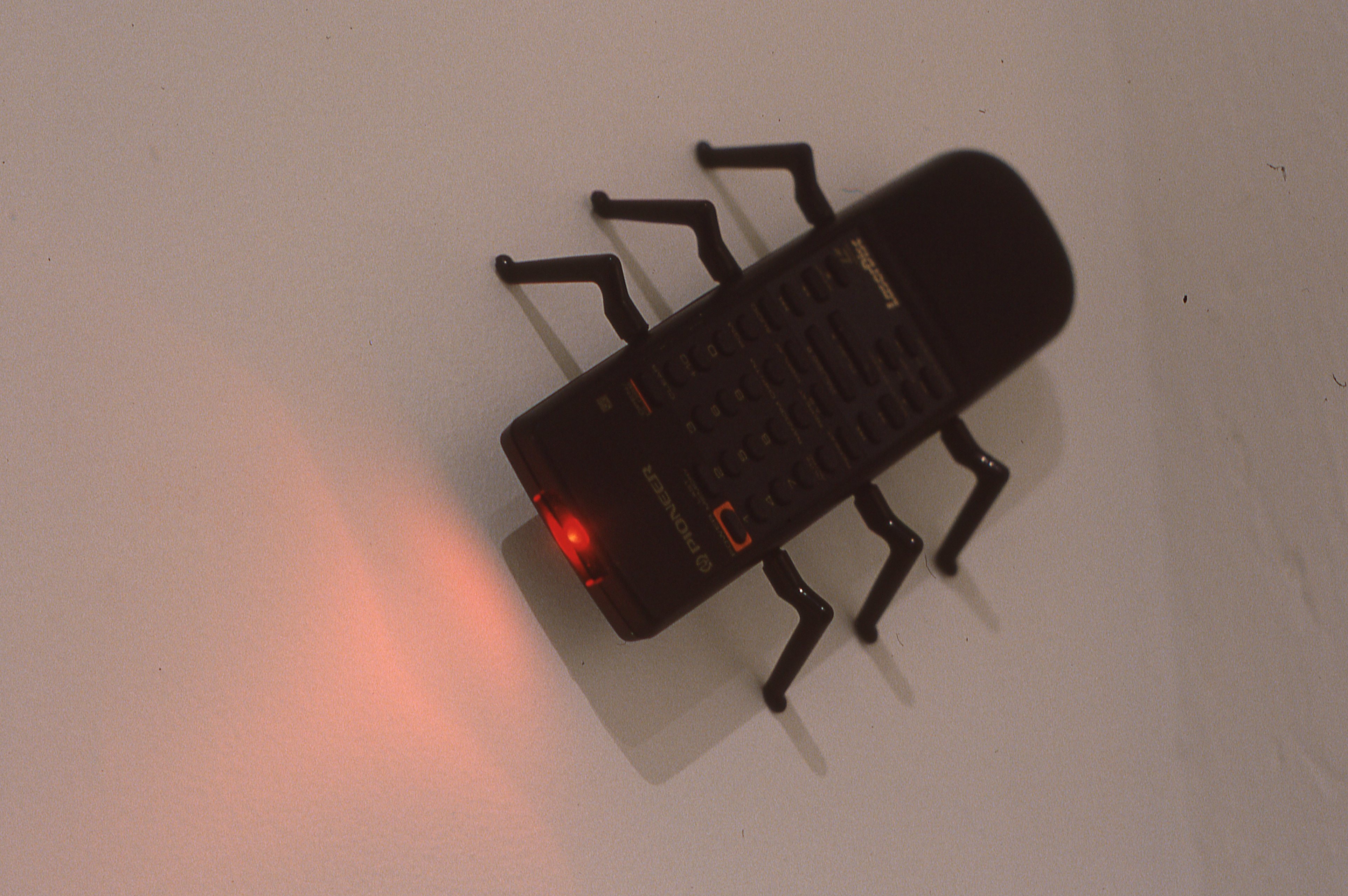 A black remote control with a red light pointing out of its head. Six insect-like legs are attached to the body of the remote, three on each side, and it is poised on the wall as if crawling.