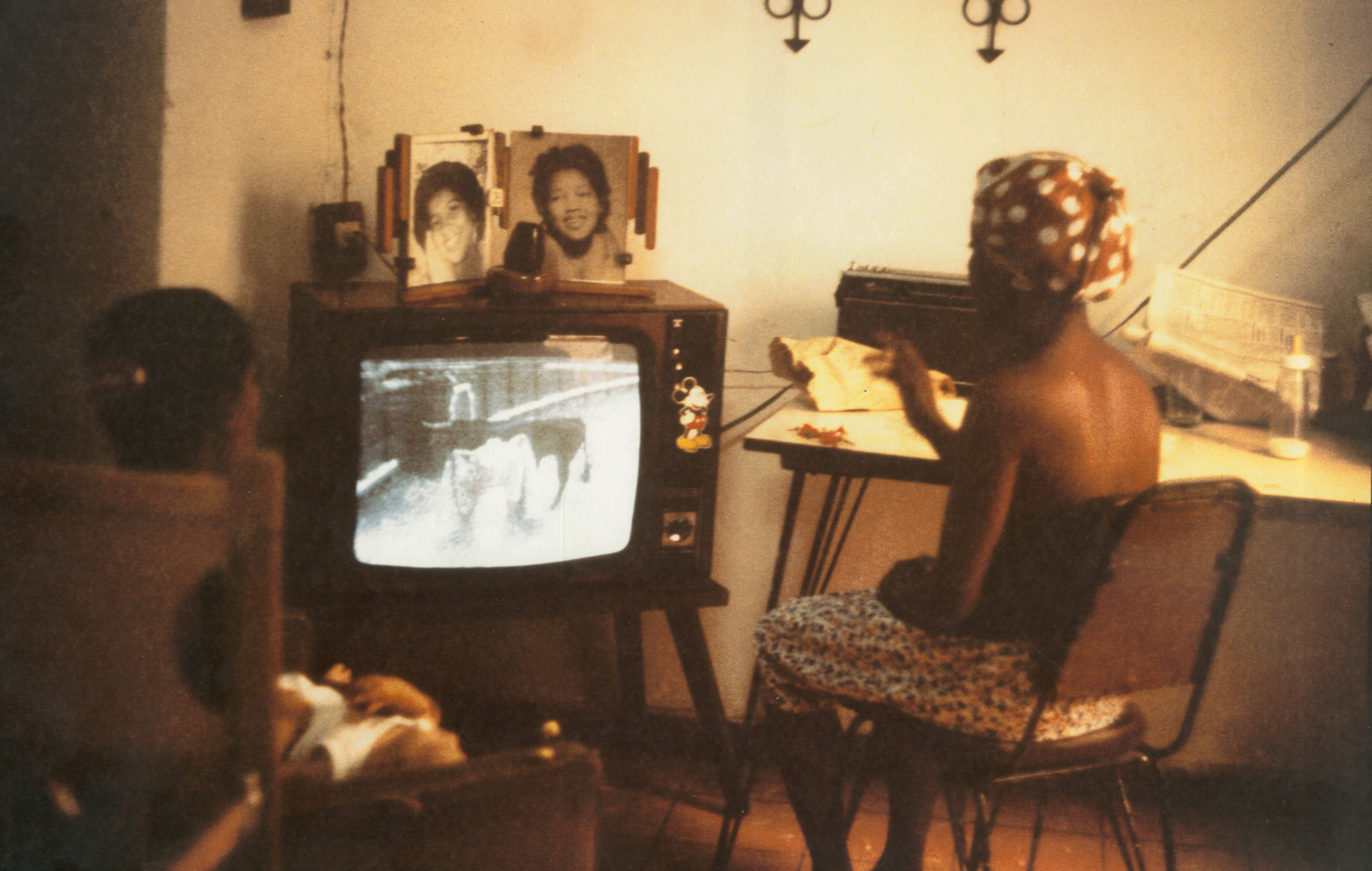 A color photographic picturing two figures who sit watching a television screen, their backs facing the camera.