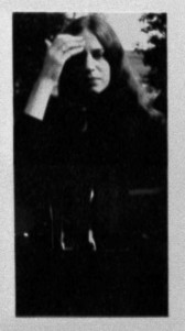 A black and white photograph of a figure with long hair. They raise their left hand up to brush away their hair from their face.