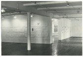 Black and white installation view of a gallery space with a large pole on the left. Several works hang from the walls.